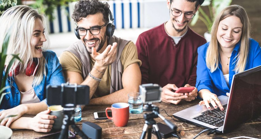 Four content marketers are having a collaborative outdoor meeting around a wooden table discussing content marketing. One person is typing on a laptop, another is using a smartphone, while the other two are in conversation. There is a red coffee mug and two cameras on tripods on the table, suggesting a creative or tech-oriented content marketing discussion.