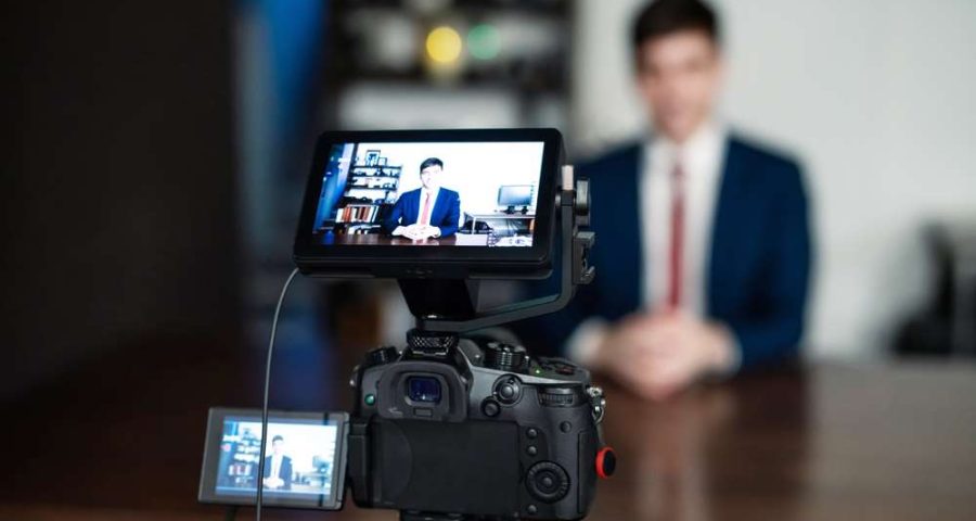 A closeup of a video camera recording a man in a suit and tie sitting at a brown desk. The man and desk are blurred in the background, so only the camera can be seen. This is supposed to represent recording a YouTube video.
