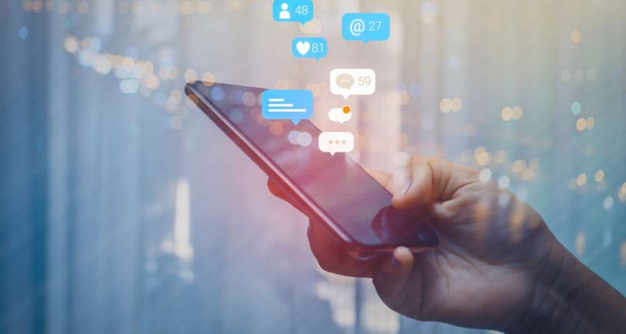 A close up of a person's hand holding a smartphone. Several thought bubbles with social media numbers can be seen above the phone screen. These are meant to symbolize social media impressions.