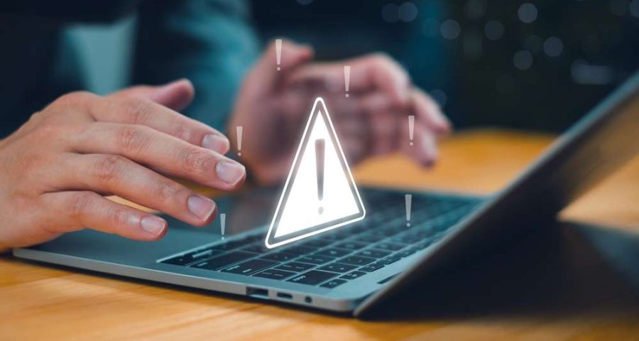 A person typing at a laptop with an illustration of a caution sign above the keyboard, meant to signify the presence of scams or spam