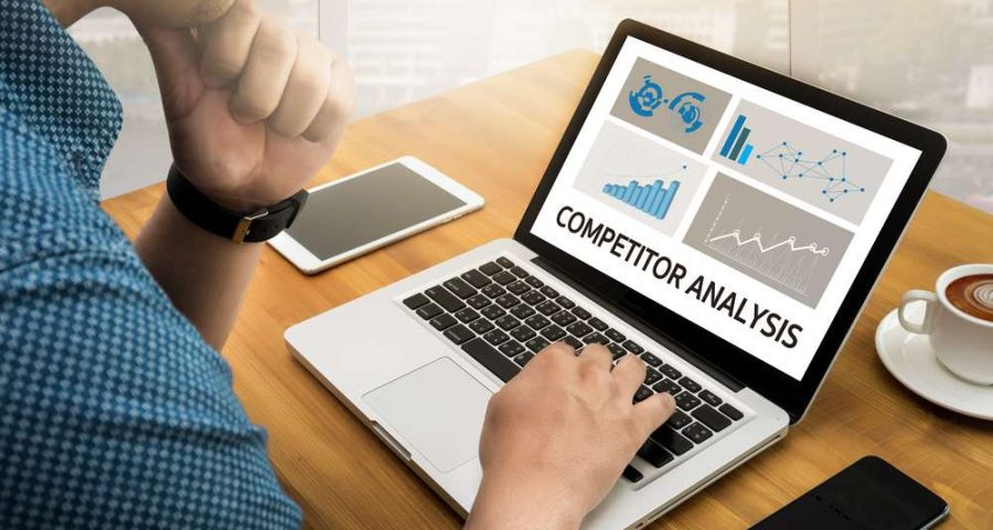 A man with a computer types on his keyboard to perform competitor analysis, as seen on the monitor