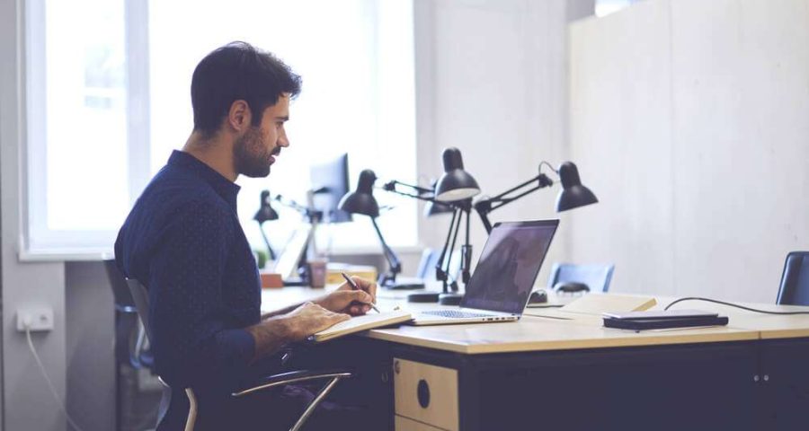 Focused male worker sits in front of laptop while writing in a notebook.